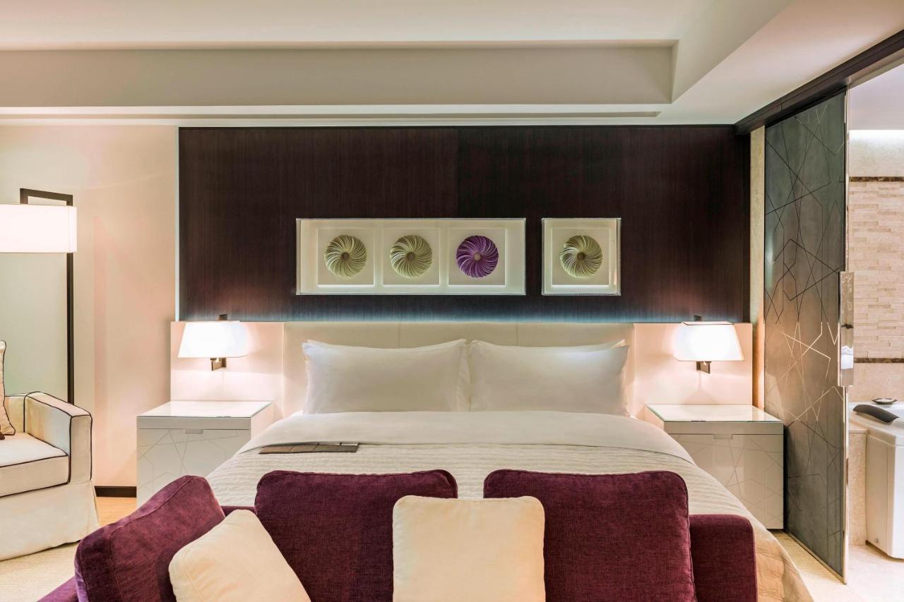 Le Meridien Dubai Hotel & Conference Centre Bagian luar foto The photo depicts a modern hotel room featuring a large, neatly made bed with white linens and a purple throw. On the wall above the bed, there are three framed artworks displayed, with a decorative element in the center. On either side of the bed, t